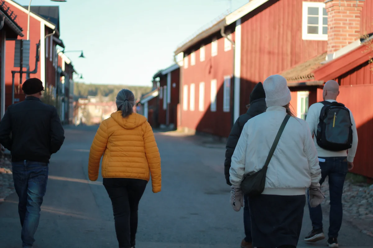 A small group gathering on a gravel path near Falun red wooden buildings at dusk