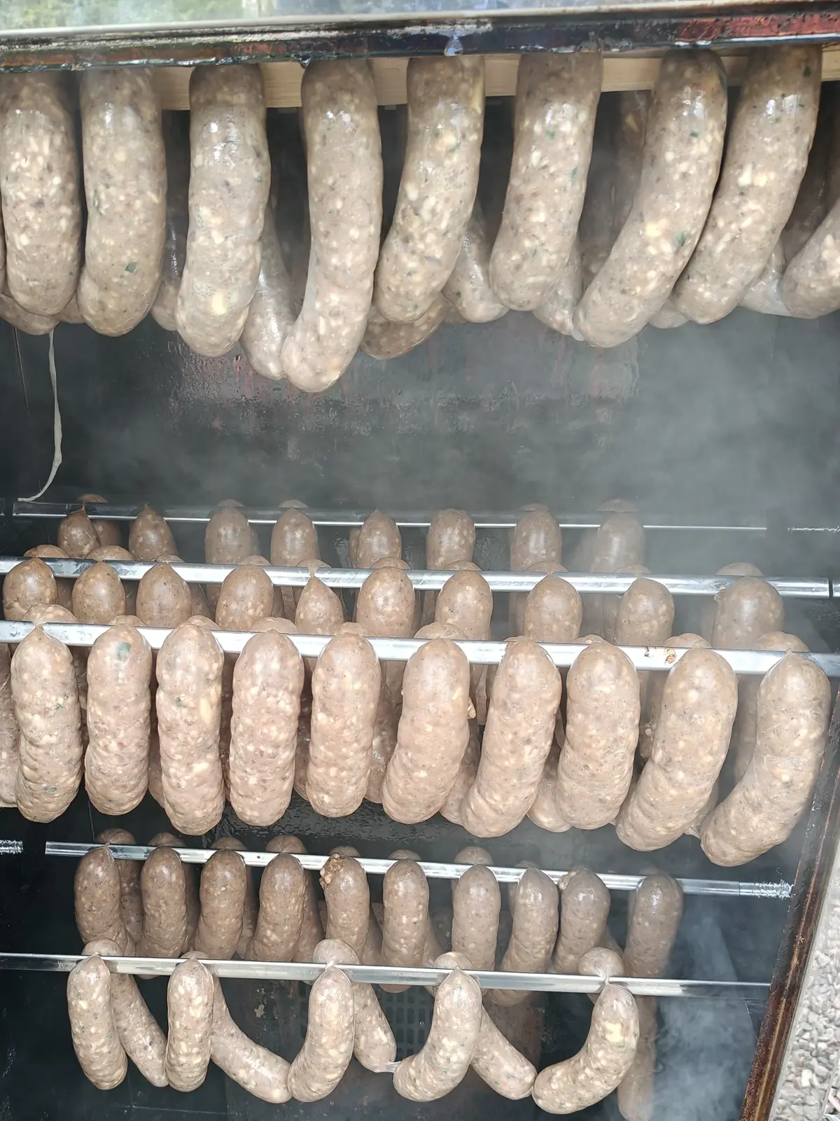 Rows of pale sausages hanging on racks inside a smoker, smoke drifting between them